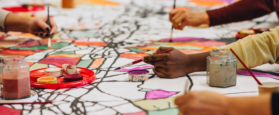 Närbild på fyra händer tillhörande olika personer håller i penslar och målar på en gemensam abstrakt duk. Måleritillbehör står också på duken. Fotografi. 