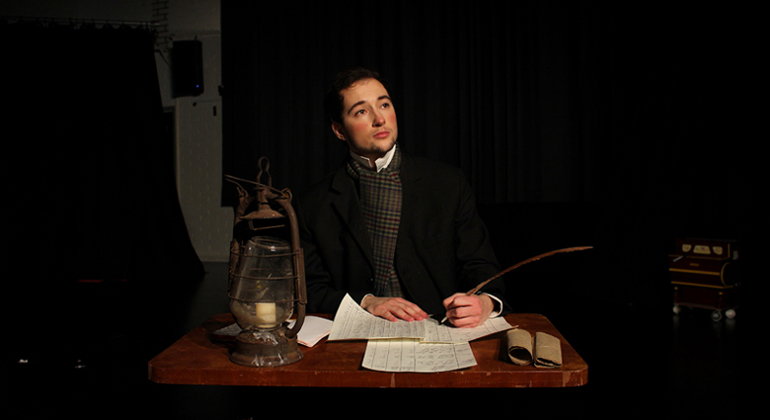 En person i historisk kl&auml;dsel skriver med fj&auml;derpenna vid ett tr&auml;bord upplyst av en lykta.