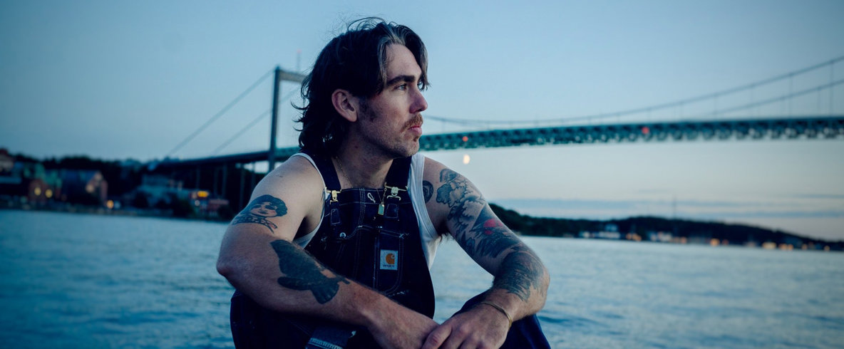 Valter Nilsson. En person med tatueringar sitter vid vattnet vid skymning. Bro i bakgrunden.