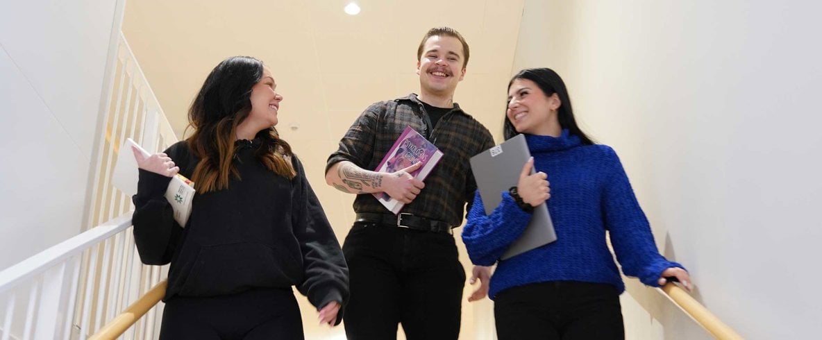 Två kvinnor och en man ler när de går nedför trappa hållande dator, böcker och studiematerial i händerna.