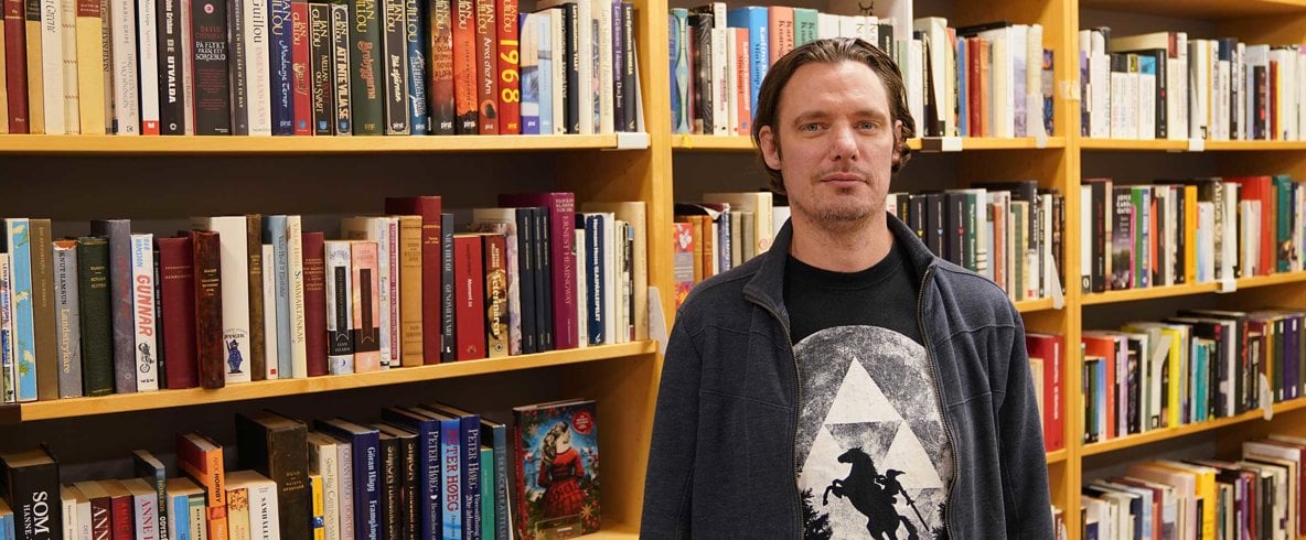 Man med mörkt bakåtkammat hår, t-shirt med tryck under mörk kofta står framför bokhylla i bibliotek.