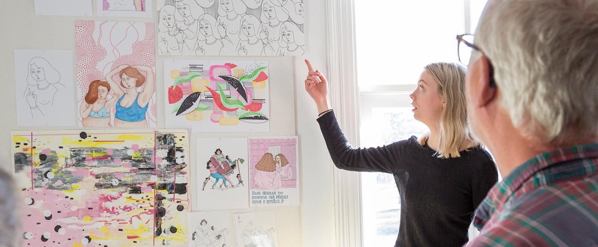 Ateljésamtal. En deltagare pekar på sina färgranna teckningar och illustrationer som är upphängda på en vägg.