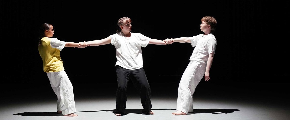 Tre personer på rad i ljuskägla i mörkt rum. Mittenpersonen håller de andra två i händerna i danspose.