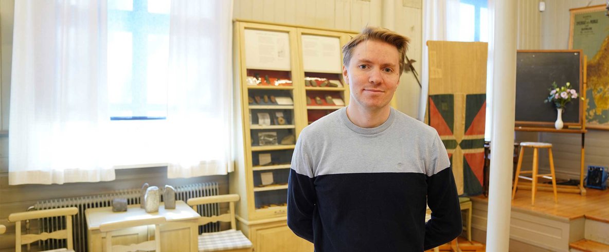 Man i kort ljusbrunt hår i halvprofil. Han har en grå och svart långärmad tröja, håller armarna bakom ryggen och ler, stående i gammaldags klassrum med fyndsamlingar och gamla kartor på väggen.