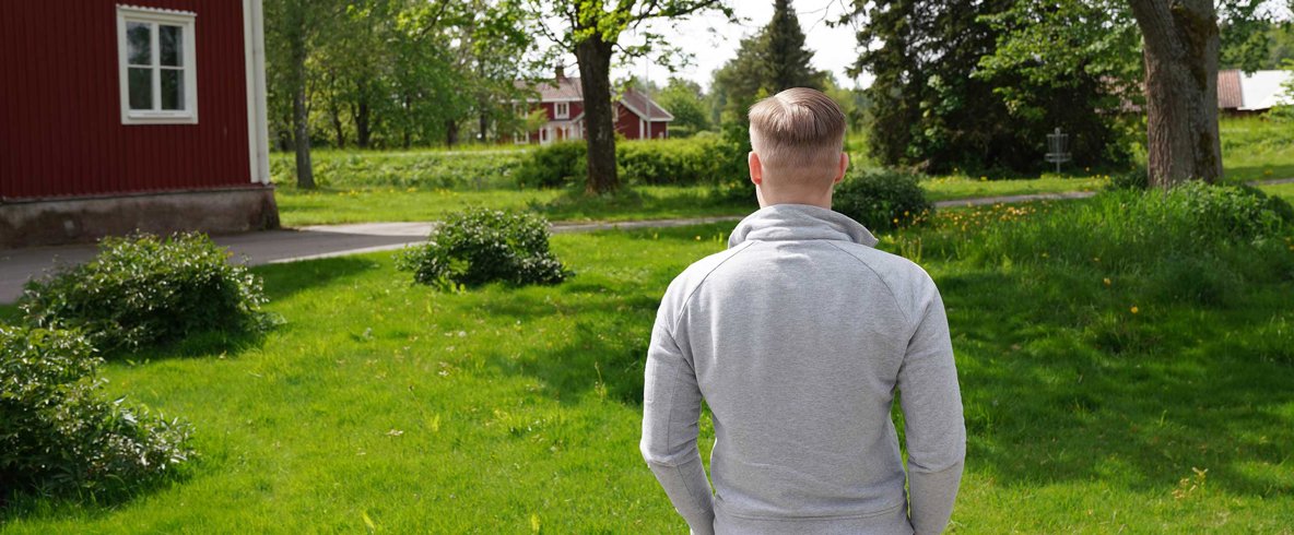 Man med kort ljust hår och grå tröja står med ryggen mot kameran och blickar ut över lantligt landskap med gröna träd och röda hus med vita knutar.