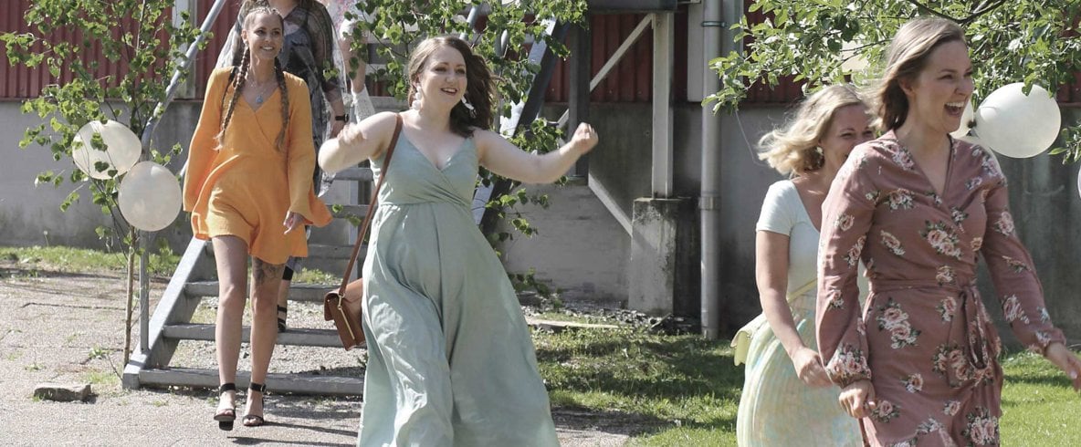 Kvinnor i festliga kläder jublar på väg ner för en trappa. Ballonger hänger i björkris.