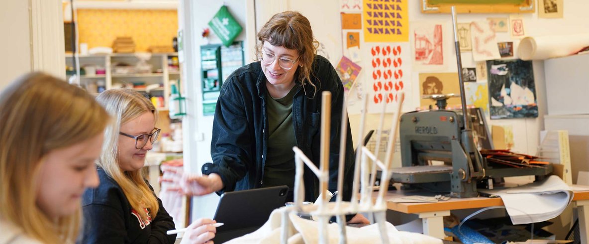 Kvinna med page, glasögon och uppknäppt mörkblå skjorta står vid bord och diskuterar med sittande kvinna som ritar med penna på skärm.