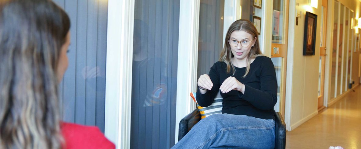 Kvinna med mellanblont långt hår, glasögon, svart långärmad skjorta och blå jeans sitter i stol och tecknar "plantera" på svenskt teckenspråk till kvinna sittande mitt emot.