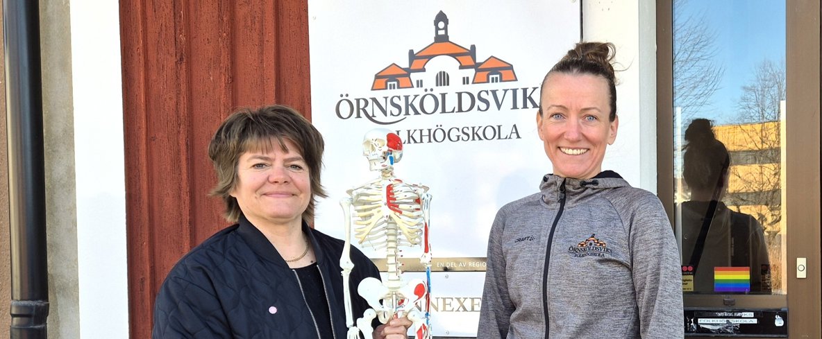 Anna Viström och Elena Dahlén håller ett litet skelett mellan sig där de står framför en skylt med folkhögskolas logga. 
