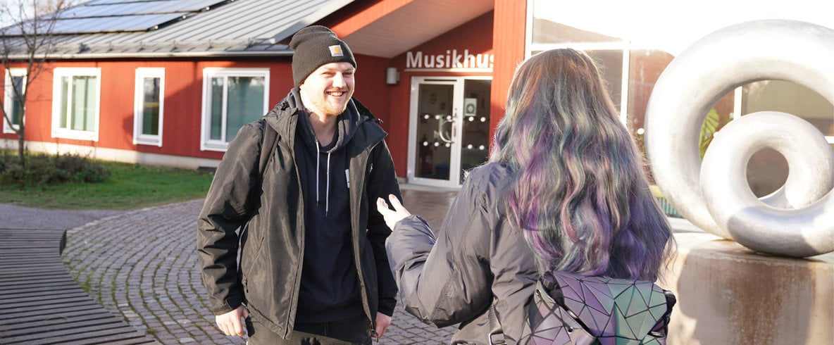 Man med mössa, svart uppknäppt jacka ovanpå blå hoodie ler och pratar med kvinna som står med ryggen till. I bakgrunden röd byggnad med texten Musikhus.