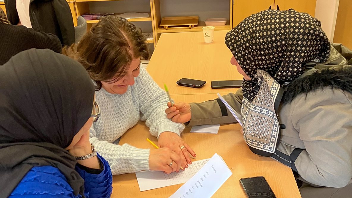 Tre kvinnor sitter vid bord i klassrum och gör uppgift med papper och penna.