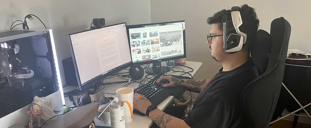 Ung man i svart t-shirt, mörkt hår och glasögon i sidoprofil. Sitter vid skrivbord med dator och dubbla bildskärmar.