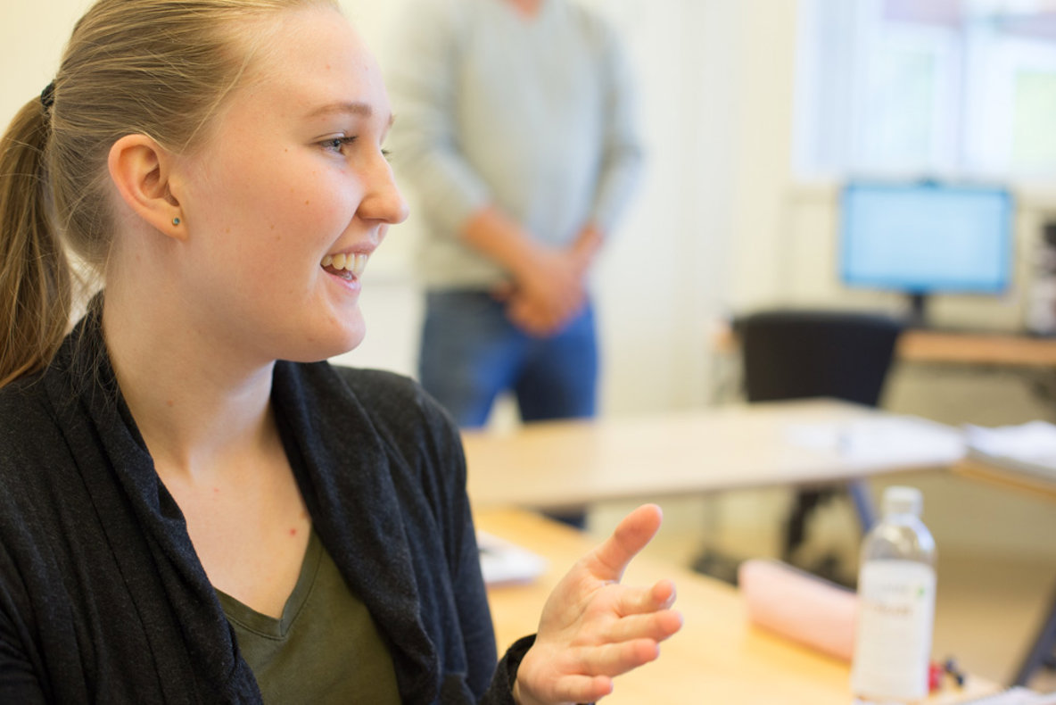 Kvinna i närbild till vänster i bild tittar till höger och håller upp vänster hand. I bakgrunden en lärare.