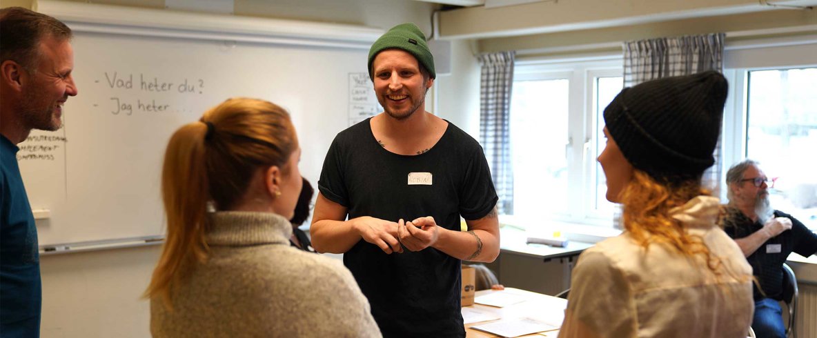 Ung man med grön mössa står i klassrum och diskuterar med två unga kvinnor.