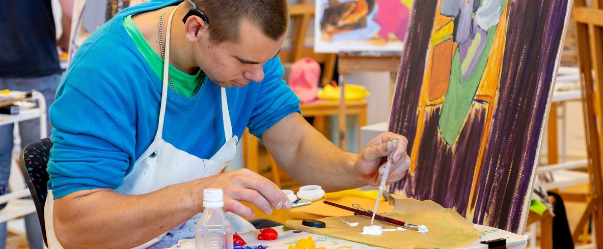 En person i blå tröja och vitt förkläde blandar målarfärg. I bakgrunden stafflin med halvfärdiga målningar. 