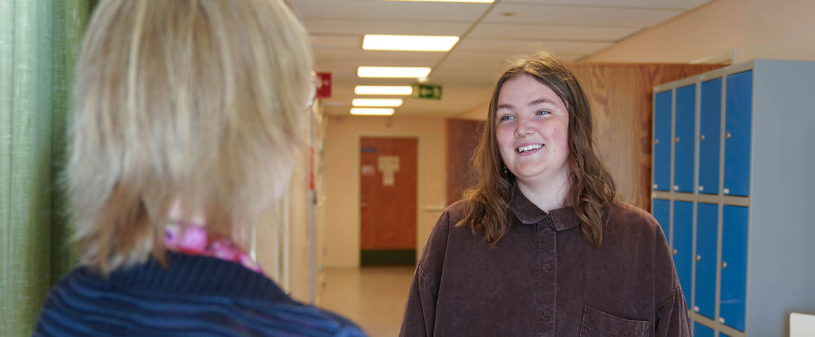 Kvinna med brunt axellångt hår och brun skjorta med armarna längs sidorna ler när hon pratar med kvinna i skolmiljö.