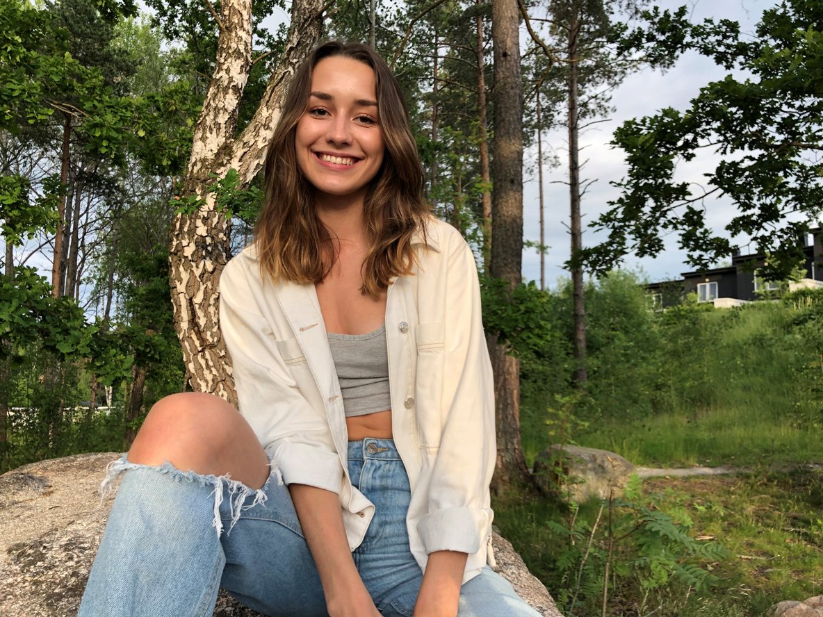 Kvinna sitter på en sten, träd i bakgrunden. Hon har mörkt vågigt hår och ler mot kameran.